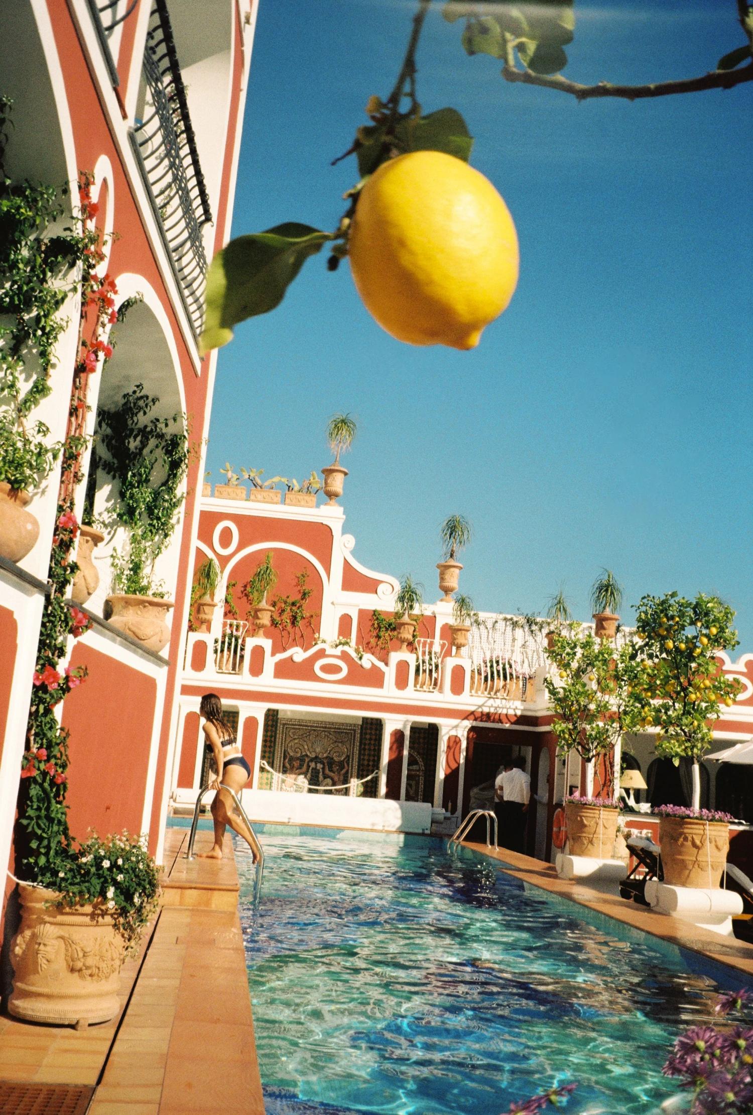 Poolside at a coral-pink villa, a lemon hanging from a branch in the foreground
