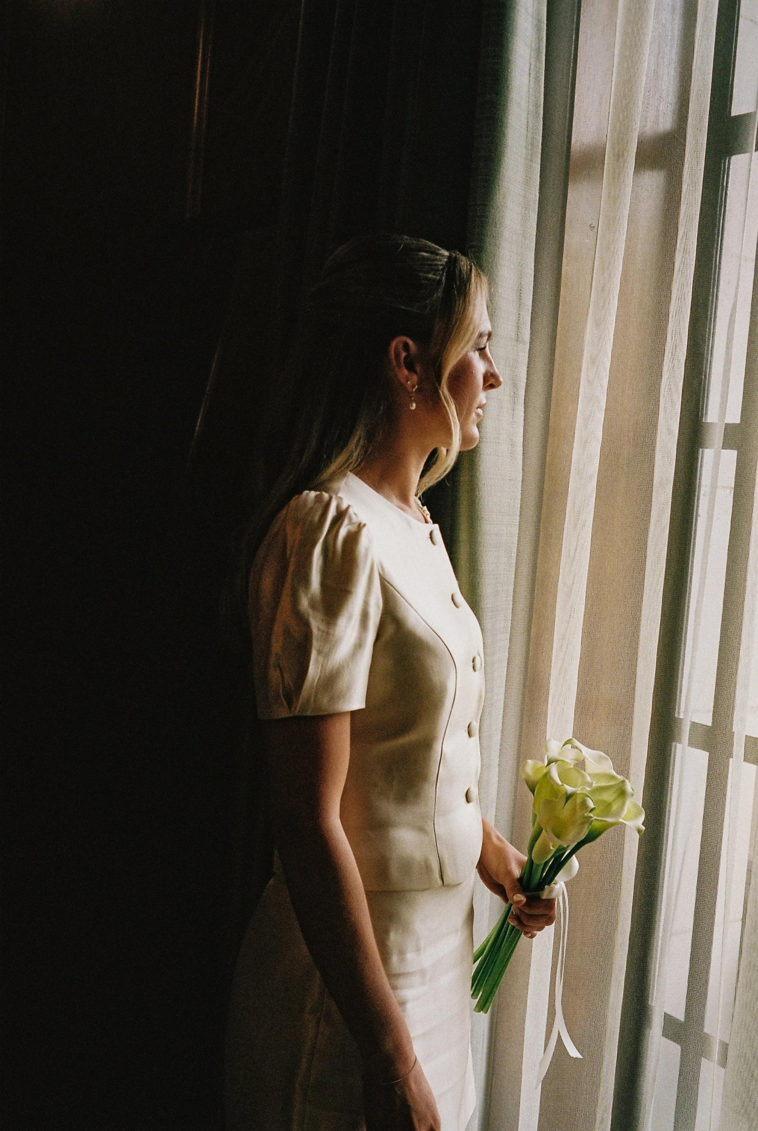 A bride in profile at a tall window, holding calla lilies, warm light against a dark interior