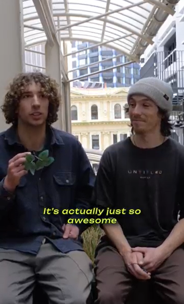 Two young men sit in front of an urban cityscape, one holding a small green leaf and the other smiling. Yellow text reads "It's actually just so awesome".