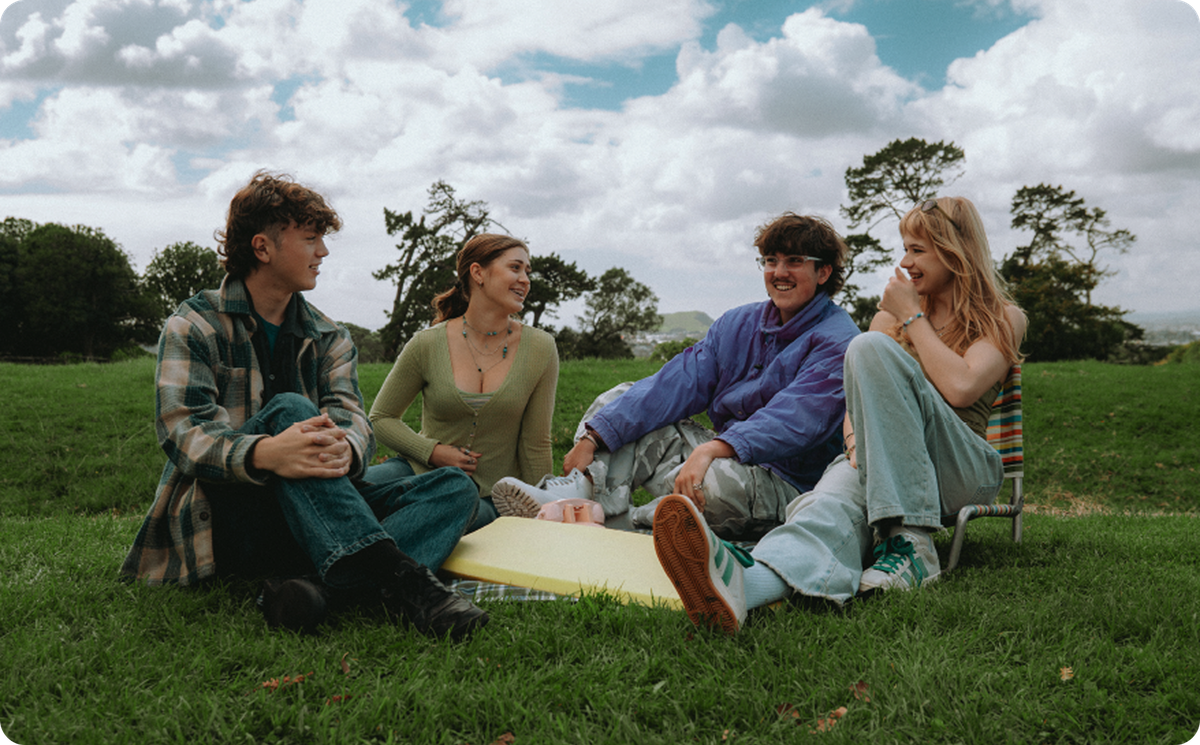 Four young people sitting in a grassy field under a cloudy sky, smiling and talking.