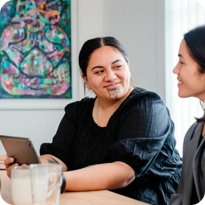 Two smiling women at a table; one has and holds a tablet.