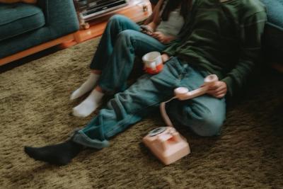 Two people sit on a green carpet, one holding a pink retro telephone receiver and a mug on their lap.