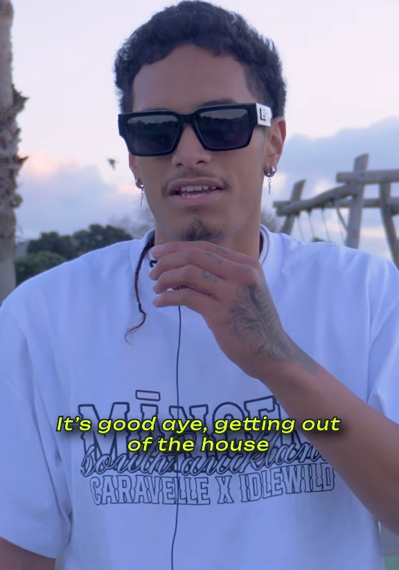 A man in sunglasses and a white graphic t-shirt with yellow subtitles that read: "It's good aye, getting out of the house."