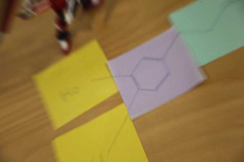 Blurry top-down view of yellow, purple, and green cards on a wooden surface, with a hexagon drawn on the purple card.