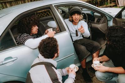 Four men with teacups gathered around a light blue car, one pointing from the driver's seat.