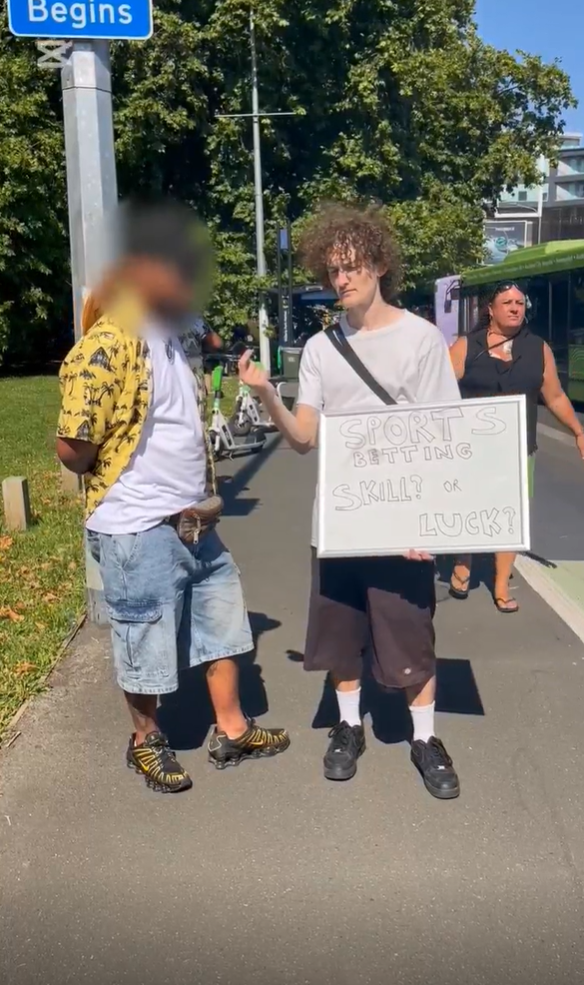 A young man with curly hair holds a sign that reads, "SPORTS BETTING SKILL? OR LUCK?"