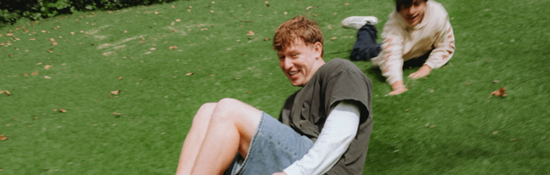 Two young men smiling as they slide and crawl down a grassy hill.