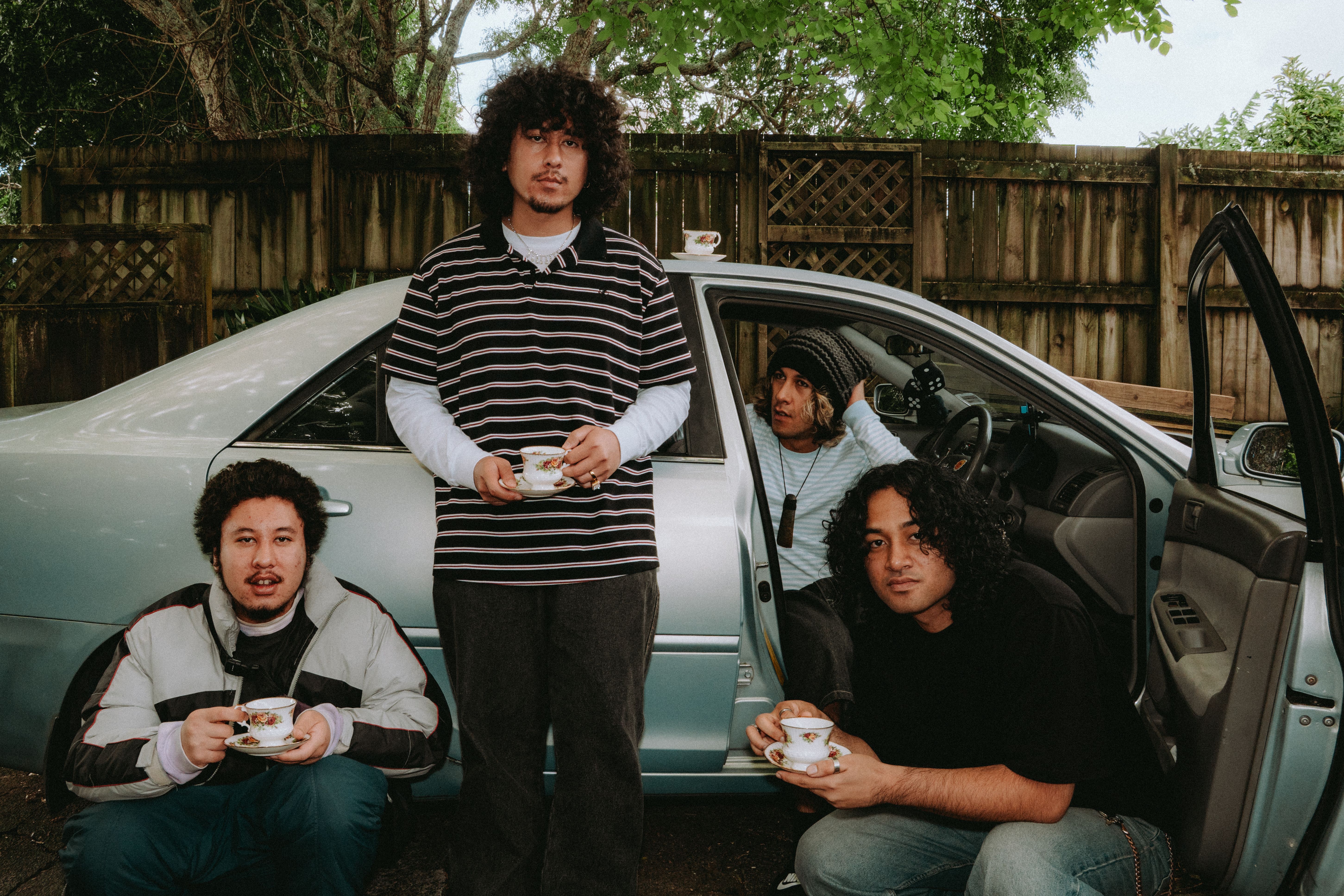 Four men holding teacups pose around a light blue car in a backyard.