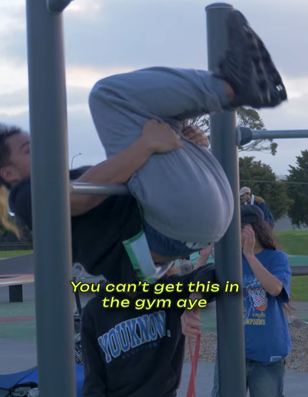 A person hangs upside down by their legs from a pull-up bar at an outdoor park. Text on the image reads, "You can't get this in the gym aye".