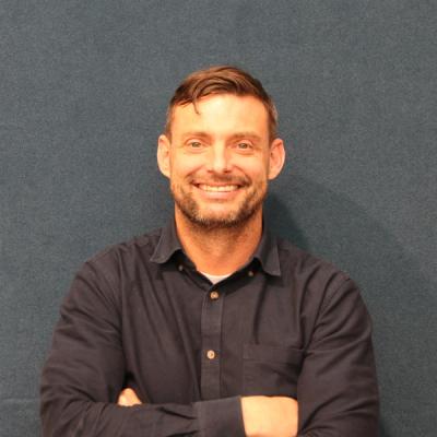 A man with a beard, wearing a navy blue shirt, smiles with his arms crossed against a dark blue background.