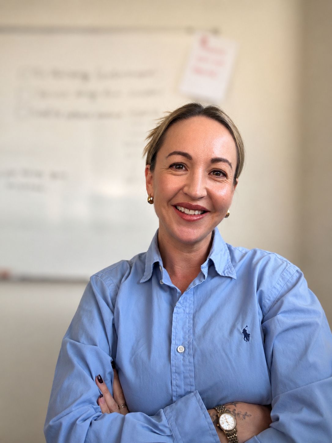 Smiling woman with light hair wearing a blue collared shirt and crossing her arms, with a blurred whiteboard in the background.