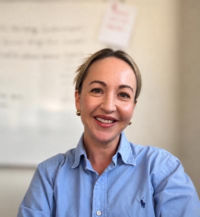 Smiling woman with light hair wearing a blue collared shirt and crossing her arms, with a blurred whiteboard in the background.