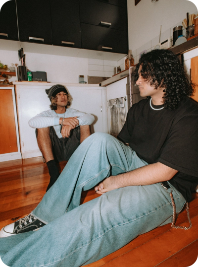 Two young men sit on a wooden kitchen floor; one with dark curly hair and a pearl necklace, the other wearing a beanie.