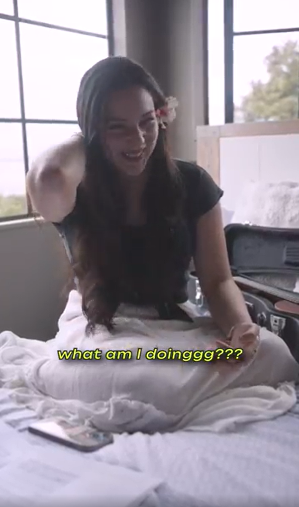 A smiling woman with a flower in her hair sits on a bed next to an open suitcase. Text reads, "what am I doinggg???".