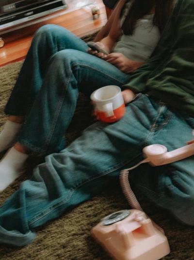 Two people sitting on a green carpet, one holding a mug and the other a phone, with a pink rotary phone in the foreground.