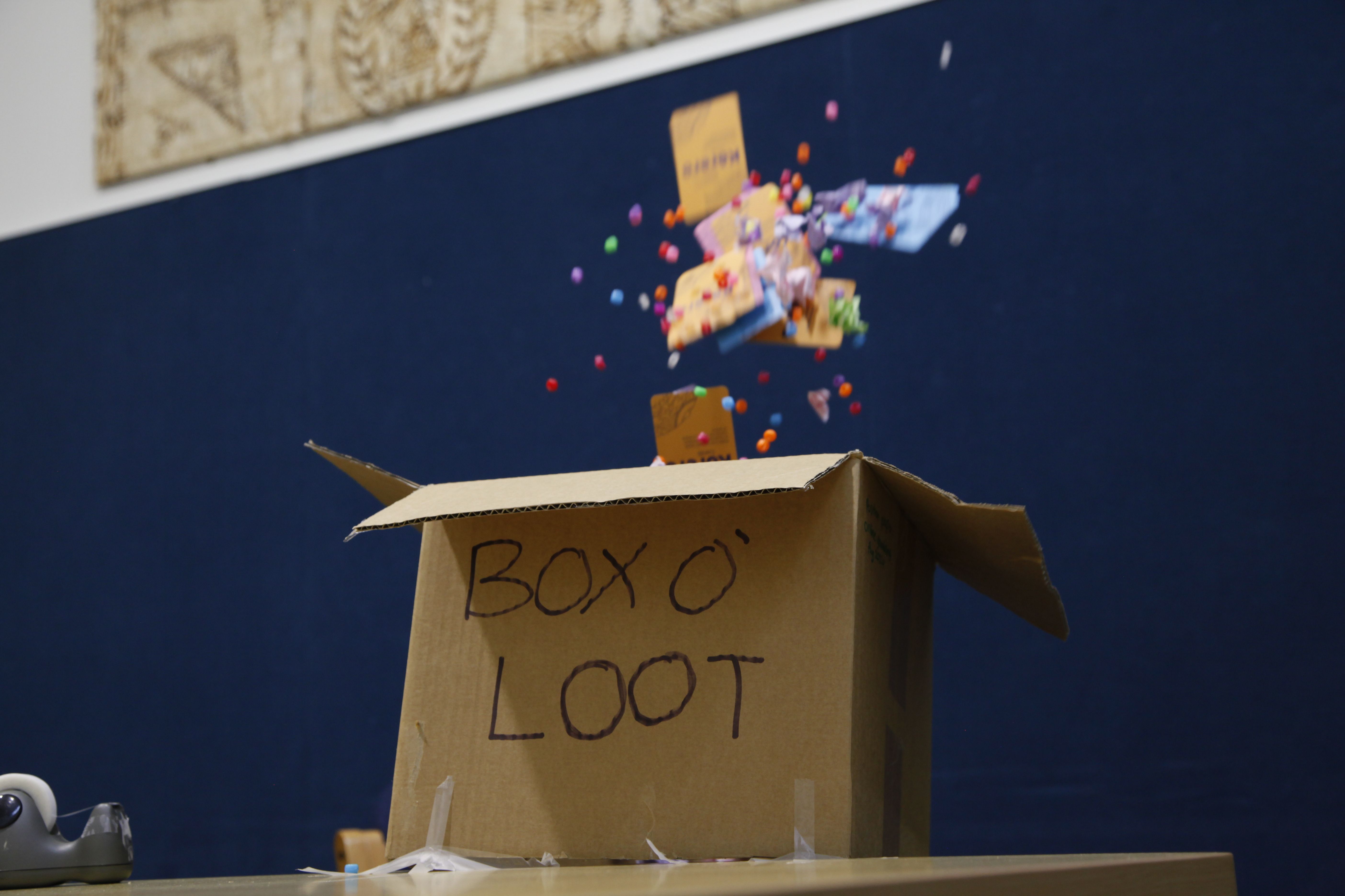 A cardboard box labeled "BOX O' LOOT" with colorful pom-poms and small items exploding upwards.