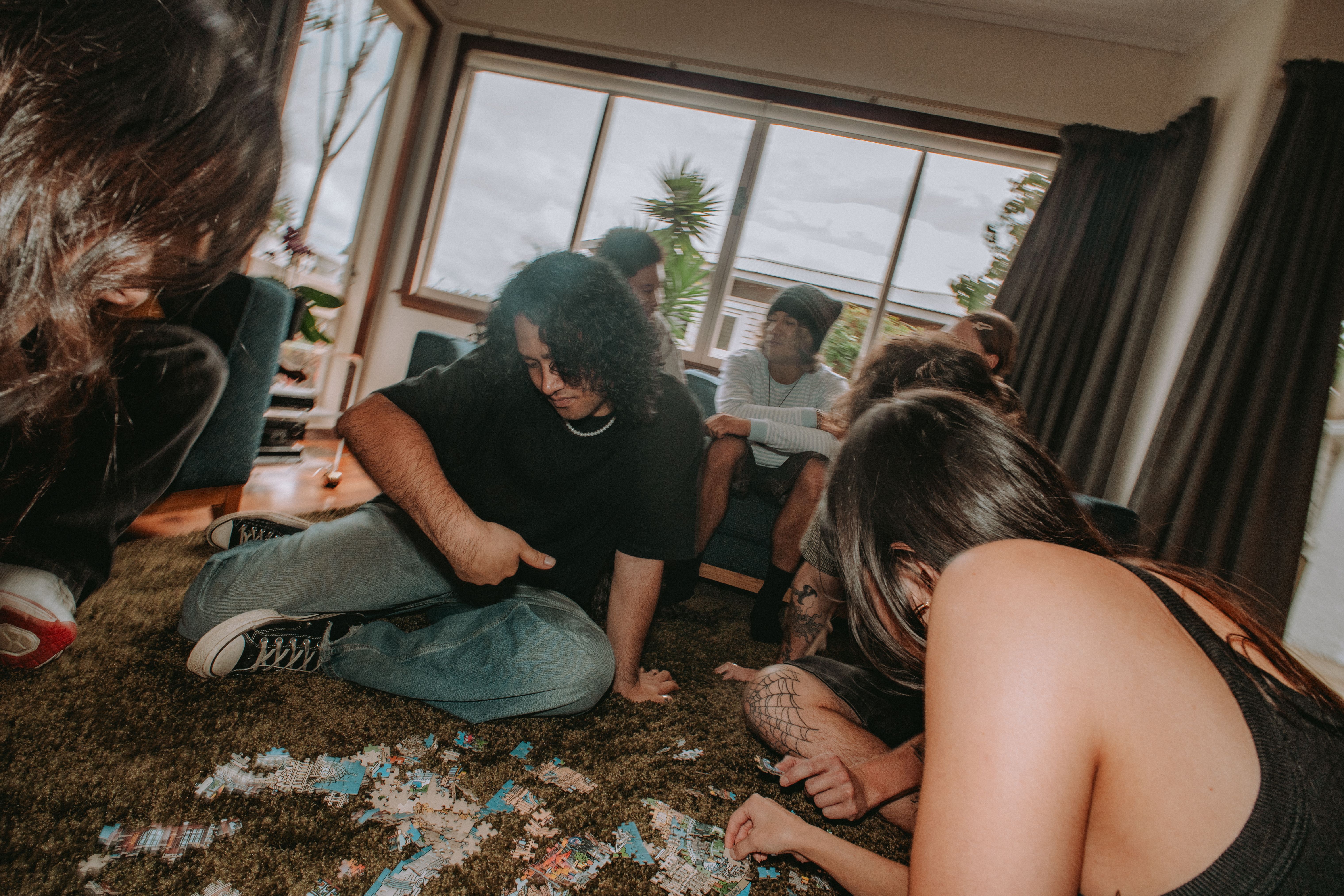 A group of young adults sits on a shag carpet in a living room, focused on a jigsaw puzzle scattered on the floor.