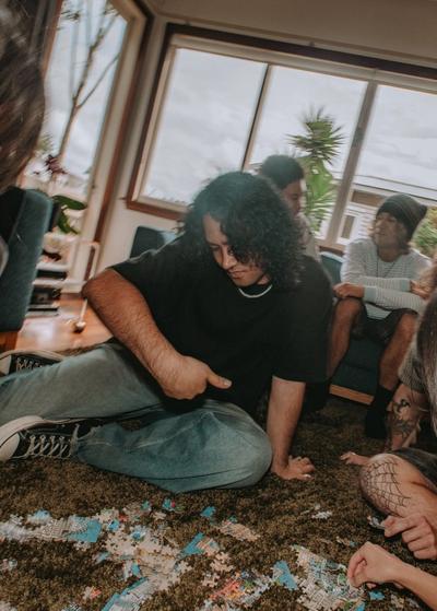 A group of young adults sits on a shag carpet in a living room, focused on a jigsaw puzzle scattered on the floor.