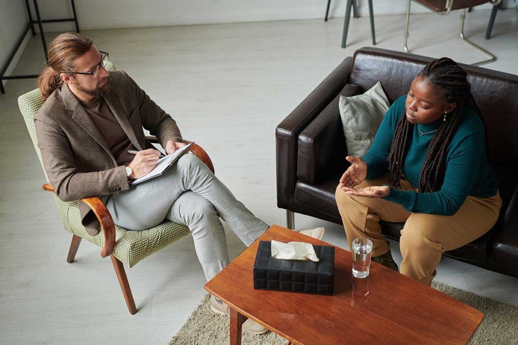 Adult client in a calm, light-filled therapy office mid-conversation, relaxed posture, warm neutral tones