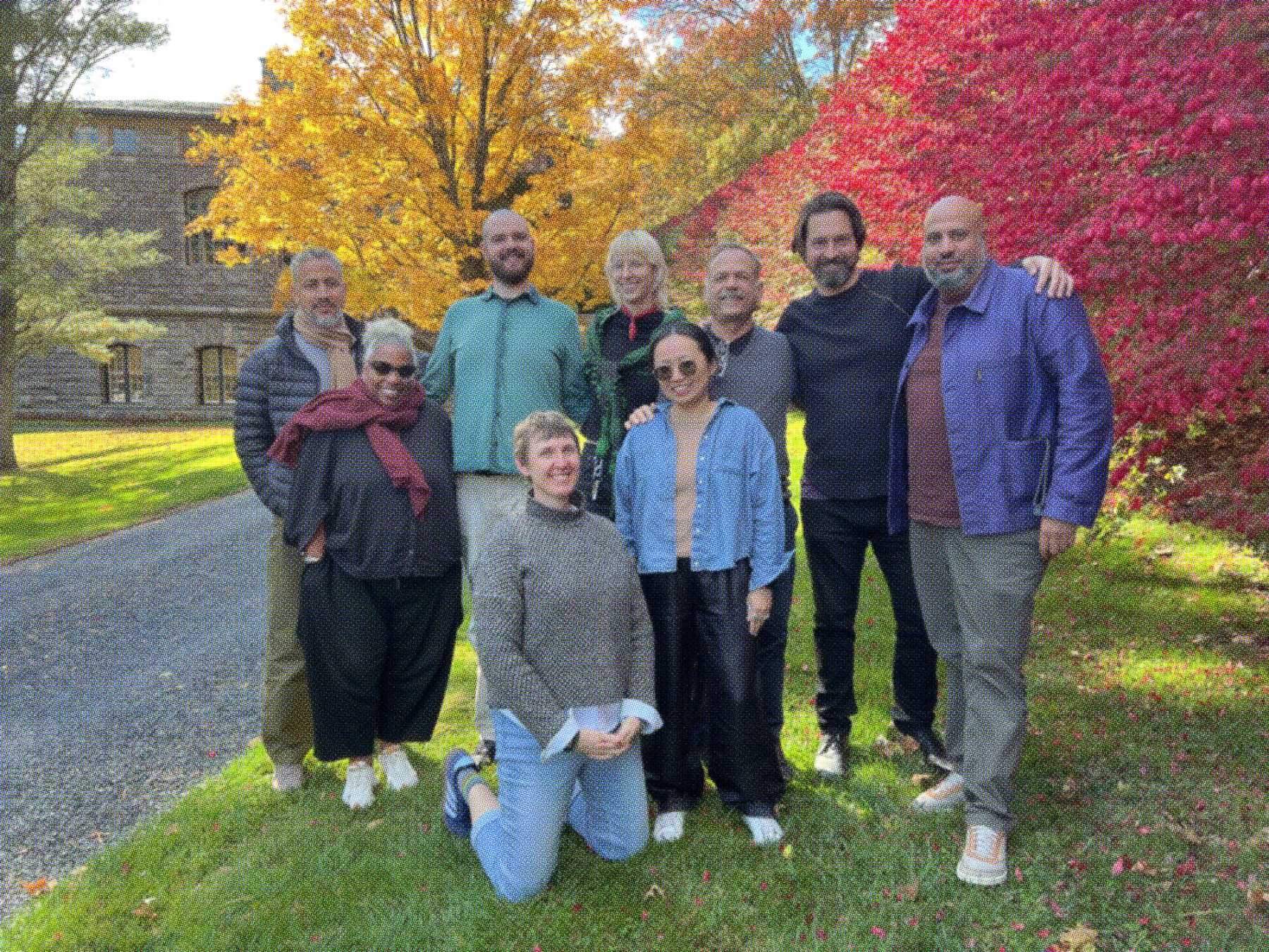 A group photo of the A Blade of Grass board and staff. Nine people pose together outside on a crisp fall day with vibrant yellow and red leaves on the trees in the background. 