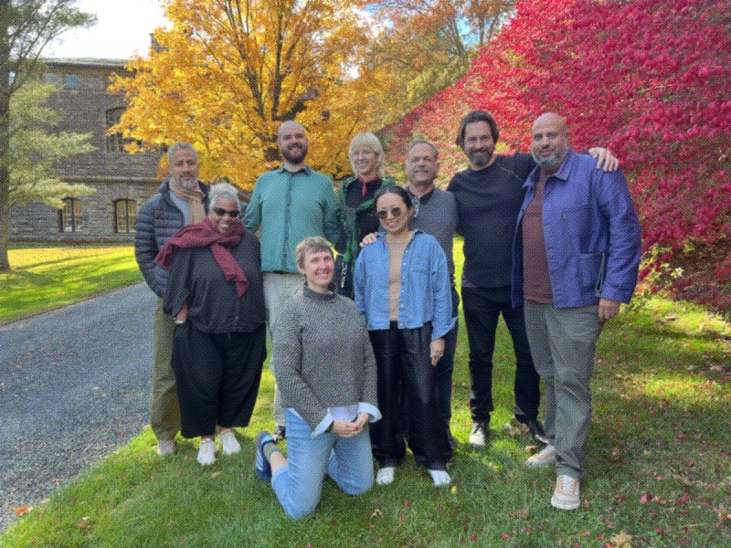 A group photo of the A Blade of Grass board and staff. Nine people pose together outside on a crisp fall day with vibrant yellow and red leaves on the trees in the background. 