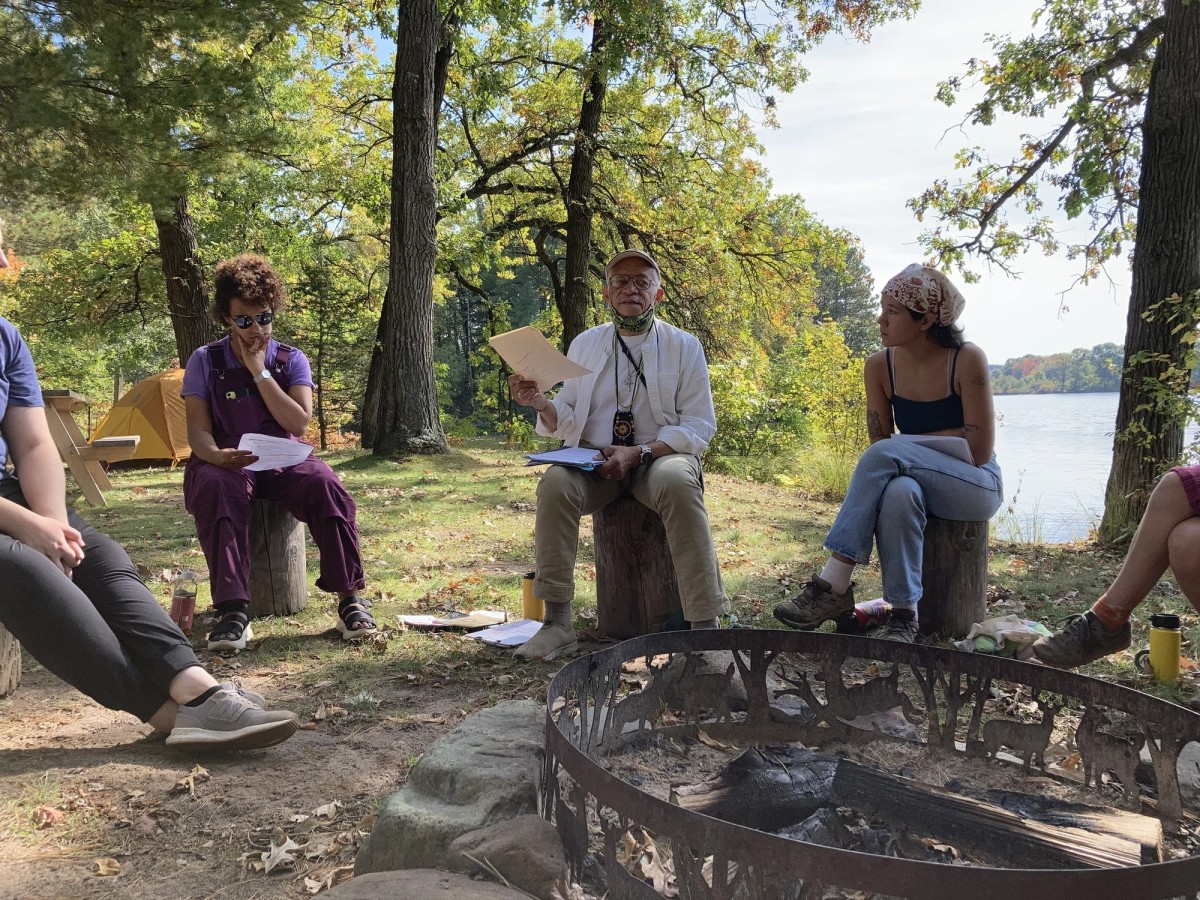 A group gathers around a firepit in the woods near a body of water during the day. 