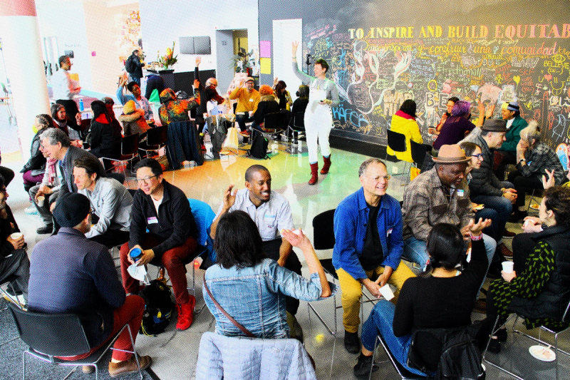 A group of artists and practitioners sit in two circles, facing each other. In the center of the circle, ashley sparks, stands with a raised arm, directing the facilitated discussion. Behind her, a text on a wall reads “To Inspire and Build Equitable…" 