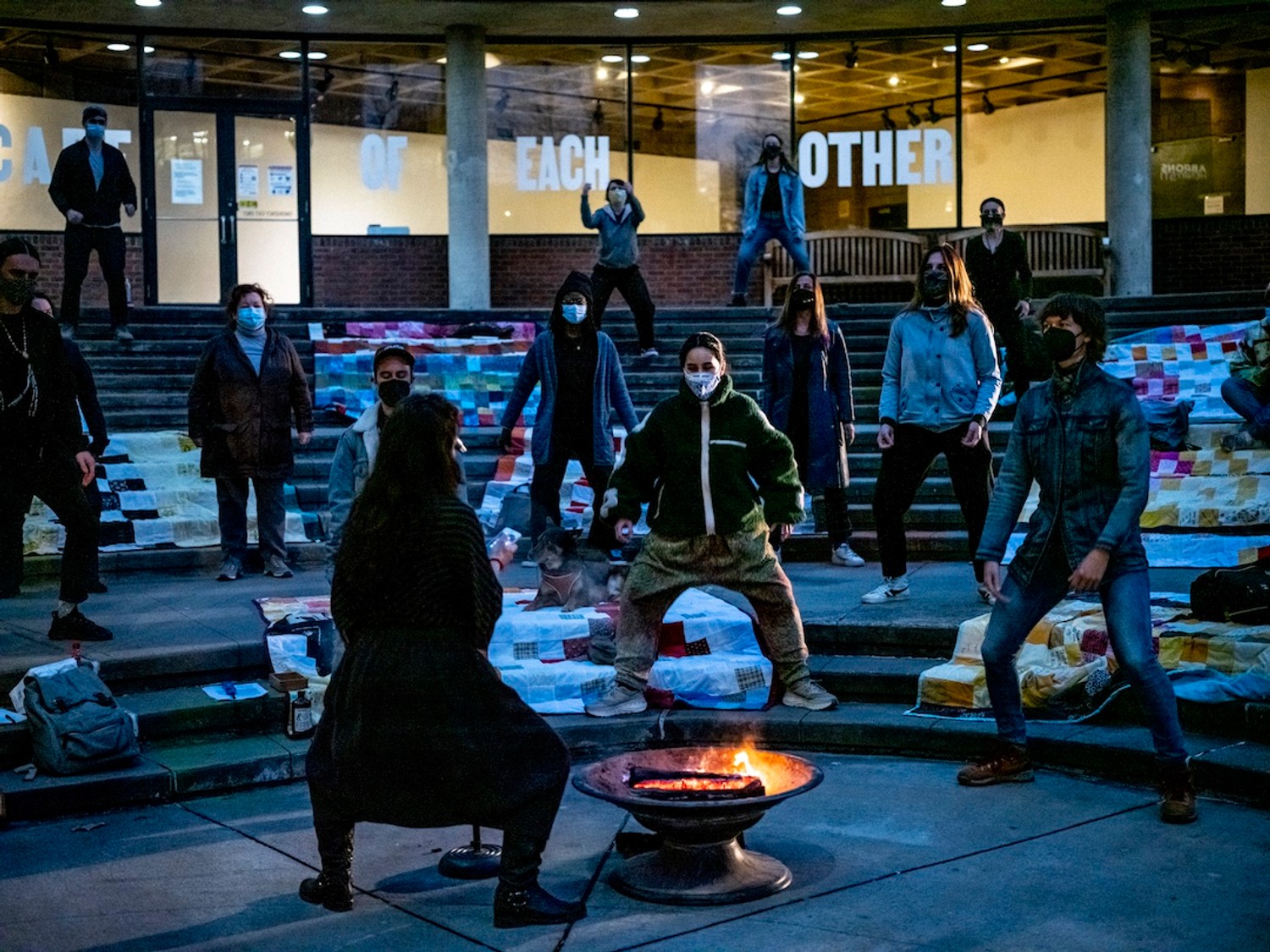 A dozen figures stand, slighting crouching and bending at the knee, along the shallow steps of a plaza in front of Abrons Art Center. They face one figure at the bottom of the shallow arena who stands next to a fire pit.