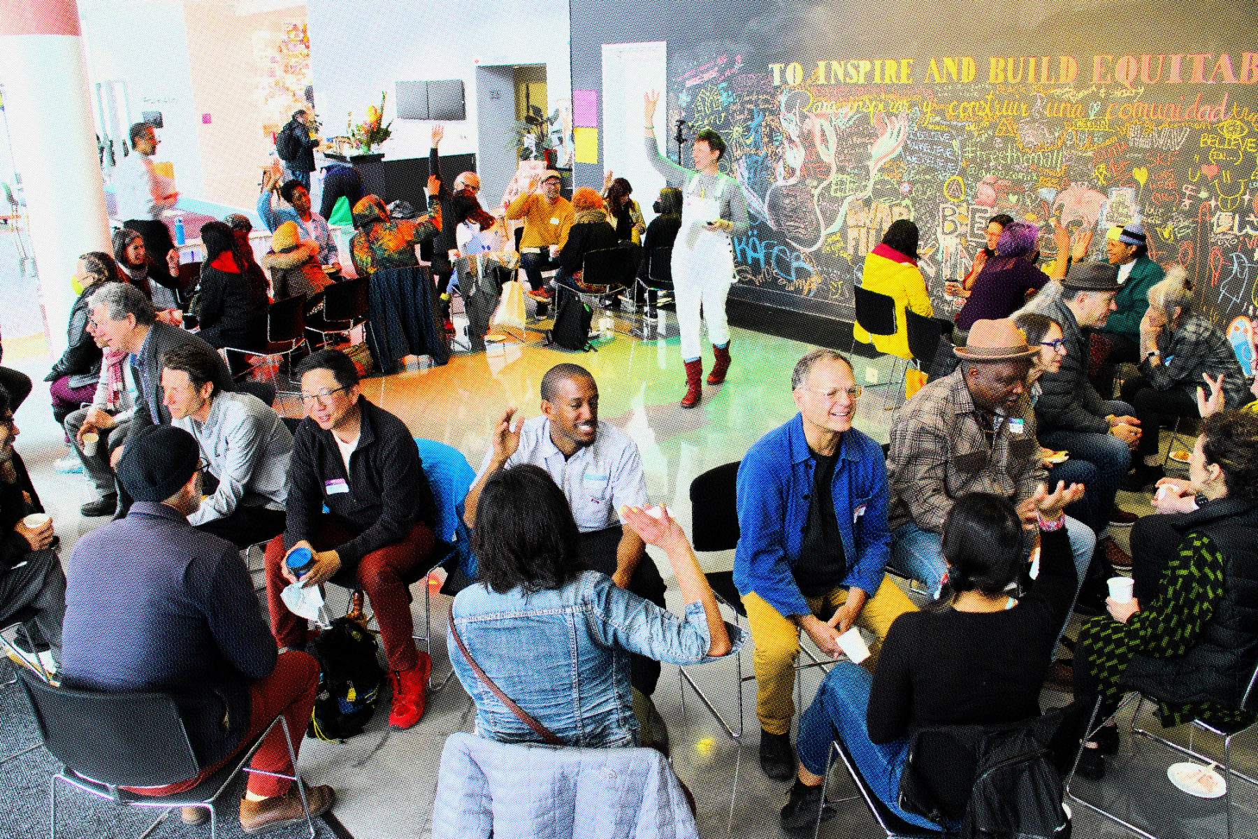 A group of artists and practitioners sit in two circles, facing each other. In the center of the circle, ashley sparks, stands with a raised arm, directing the facilitated discussion. Behind her, a text on a wall reads “To Inspire and Build Equitable…"