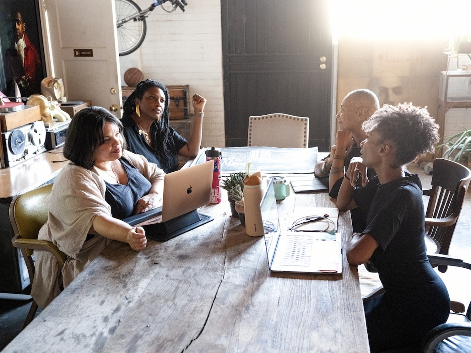 A candid photograph of a group of four African American women gathered around a table covered with papers and materials, in a working session. Daylight comes in from the left side of the frame, and the mood is informal and focused.