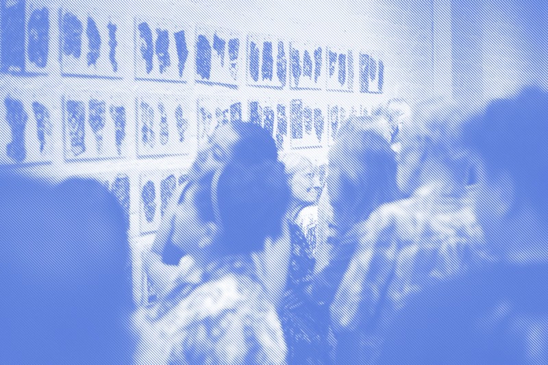 Gallery visitors closely examine a grid of small, densely patterned ink drawings mounted in rows on a white brick wall. Several people lean in to look at the work, seen from behind and in profile.
