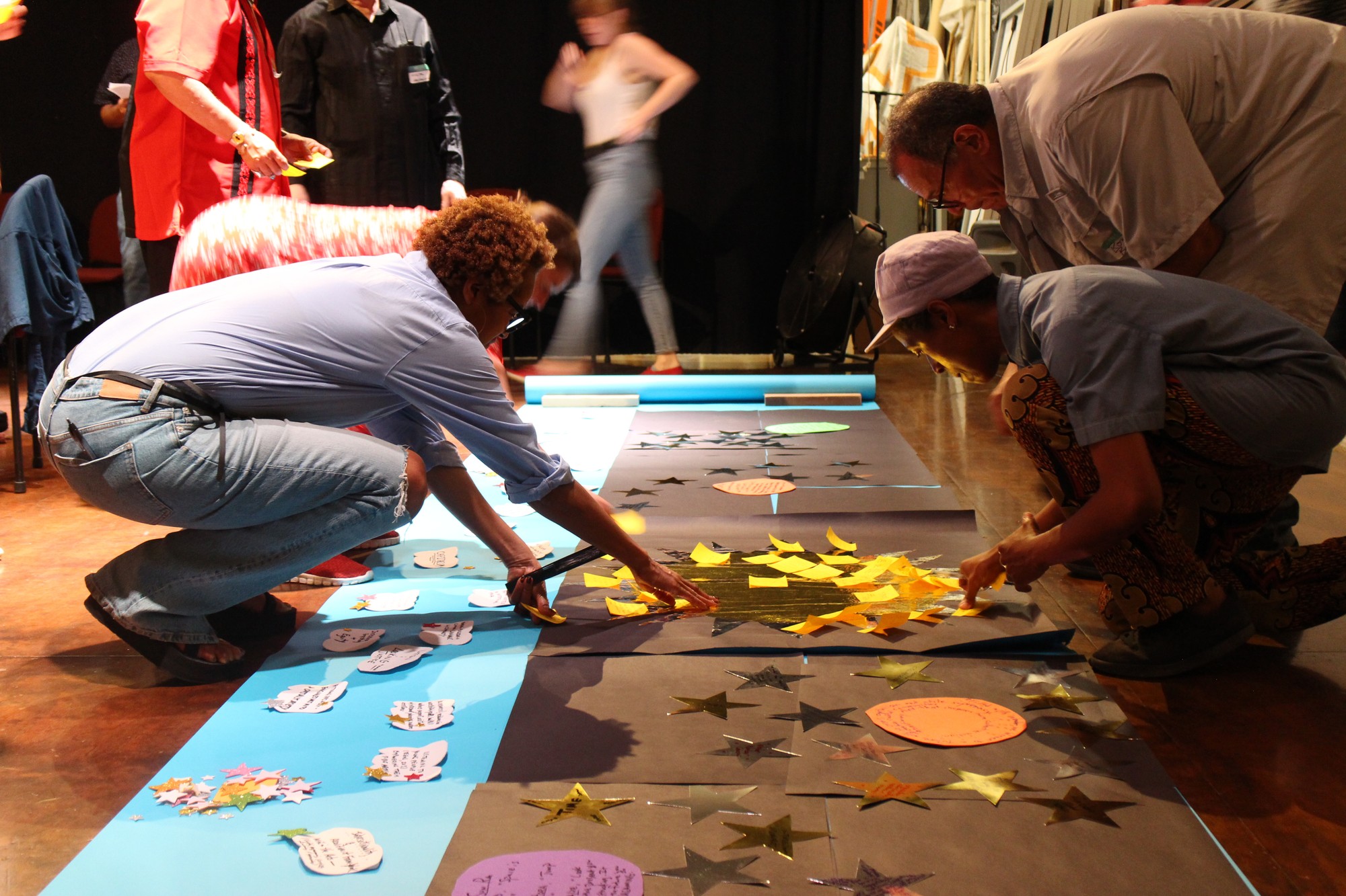 In a warmly lit theatre, several research participants crouch over a large sheet of black and blue paper, affixing stars and clouds with their notes onto the paper. 