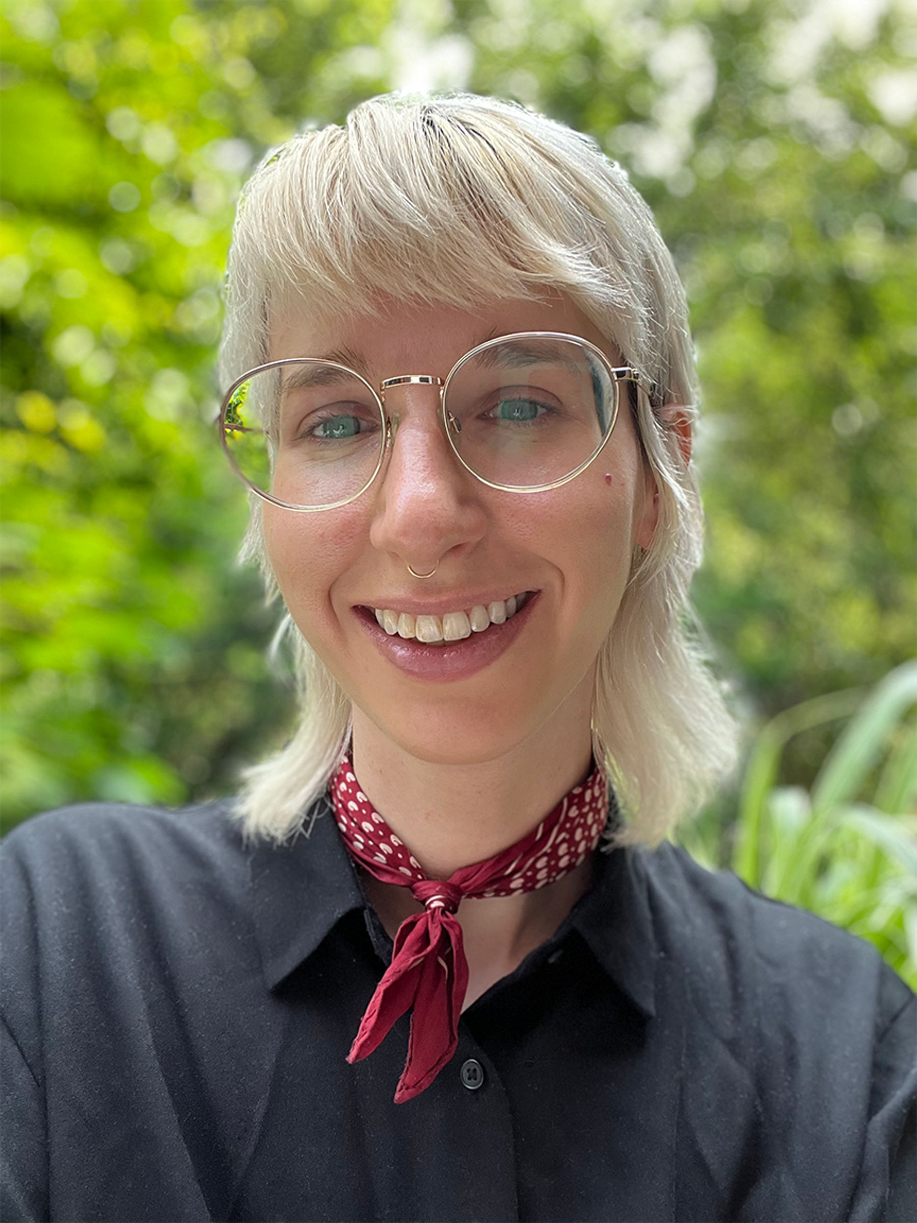 A photo of Allie, a white person with bleached blonde hair and glasses, smiles into the camera in front of a lush, green background.