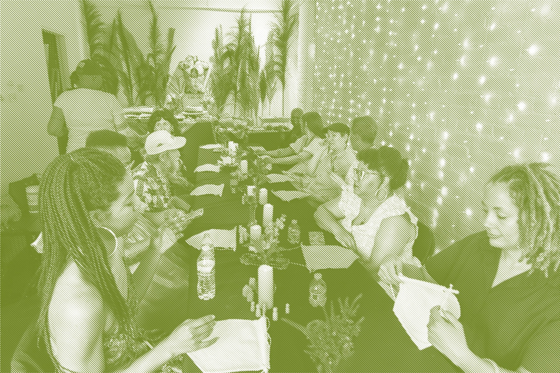A long table set with tablecloths, candles, small flower arrangements, and water bottles in a warmly lit gallery space. Guests are seated on both sides, some holding or working on small white fabric banners. The back wall is decorated with tall dried grass plumes and string lights, with a food spread visible at the far end of the table.