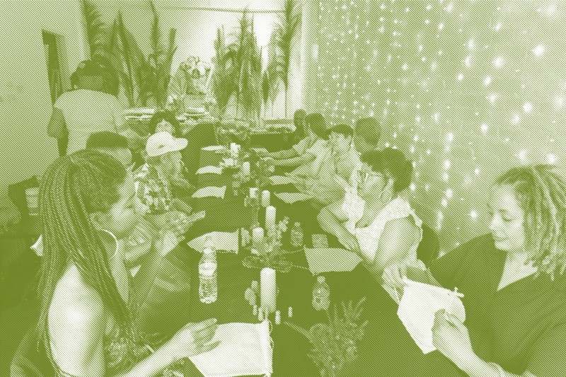A long table set with tablecloths, candles, small flower arrangements, and water bottles in a warmly lit gallery space. Guests are seated on both sides, some holding or working on small white fabric banners. The back wall is decorated with tall dried grass plumes and string lights, with a food spread visible at the far end of the table.