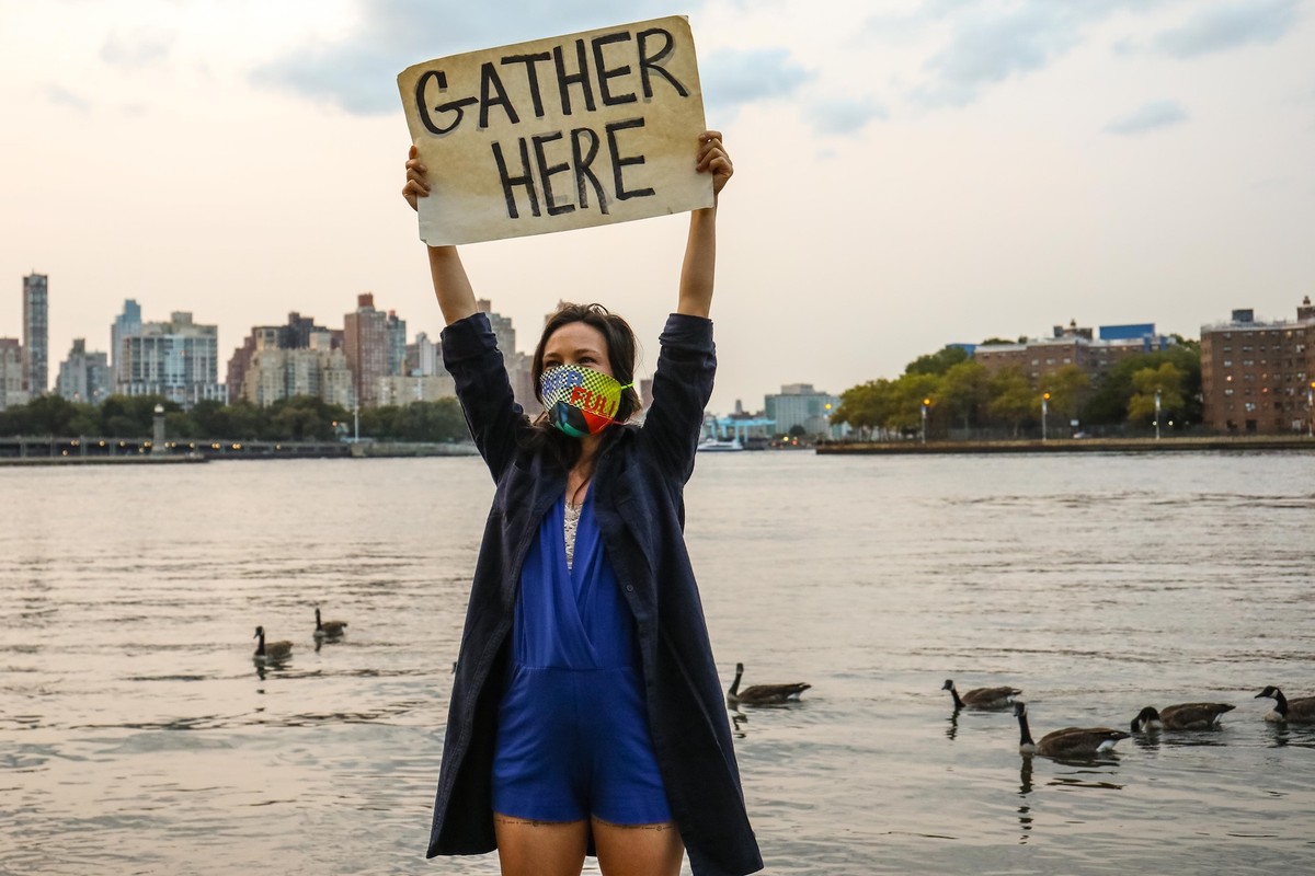 Emily Johnson, a Yup’ik woman, stands at the edge of the East River in Lenapehoking. She has long black hair, and wears a vibrant facemask designed by artist Jeffrey Gibson, with the letters POWER and FULL on it. Her arms are raised over her head and she holds up a hand-written sign that reads “GATHER HERE”. Behind her, the gray water is spotted with ducks, and some of the Manahatta skyline is visible.