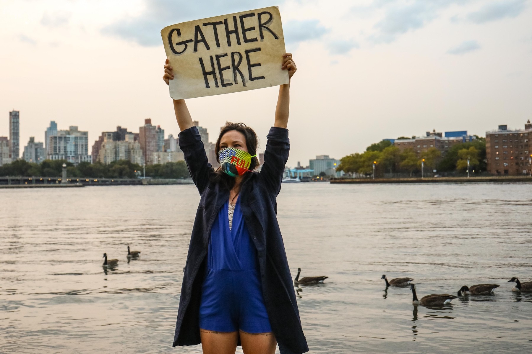Emily Johnson, a Yup’ik woman, stands at the edge of the East River in Lenapehoking. She has long black hair, and wears a vibrant facemask designed by artist Jeffrey Gibson, with the letters POWER and FULL on it. Her arms are raised over her head and she holds up a hand-written sign that reads “GATHER HERE”. Behind her, the gray water is spotted with ducks, and some of the Manahatta skyline is visible.