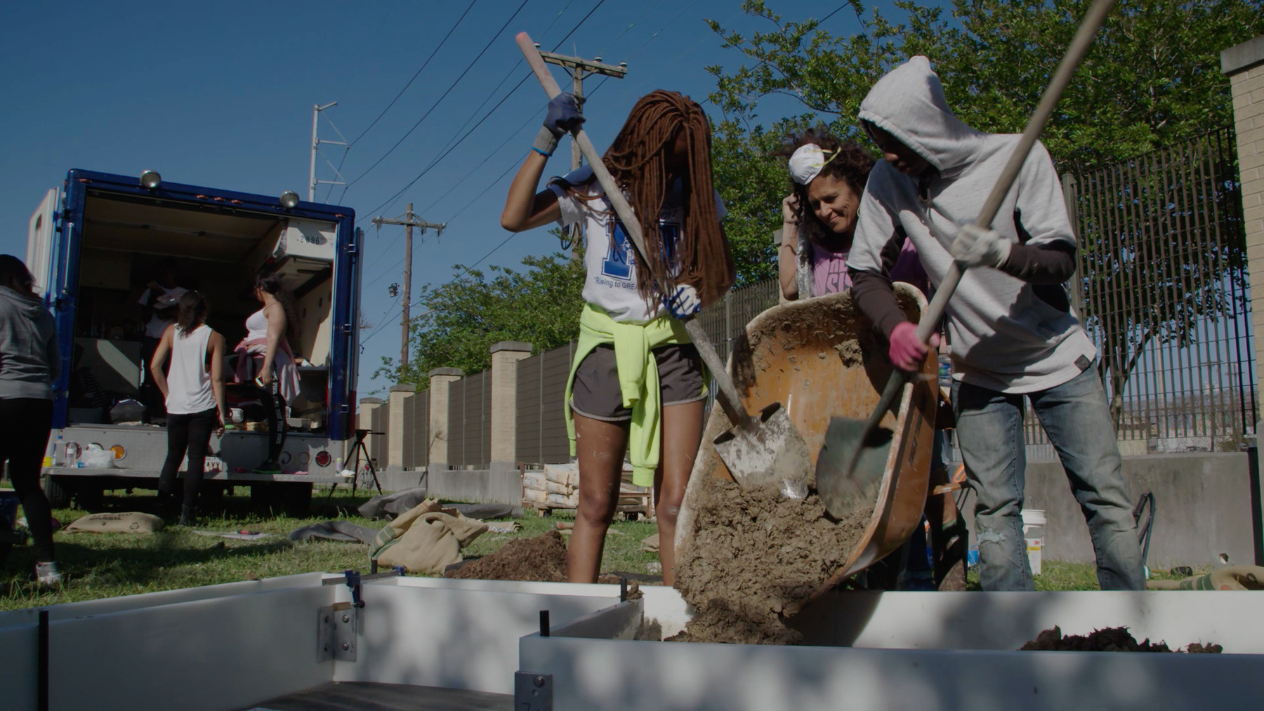 A woman stands between two young students who are shoveling dirt out of a wheelbarrow with two shovels into a garden bed made out of concrete. 