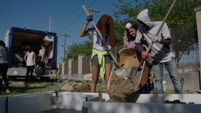 A woman stands between two young students who are shoveling dirt out of a wheelbarrow with two shovels into a garden bed made out of concrete. 