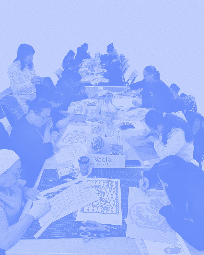 A group photo of many women at a table working on cut paper artworks. Artist Christine Wong Yap stands behind them. The photo is rendered in a periwinkle blue and white bitmap. 