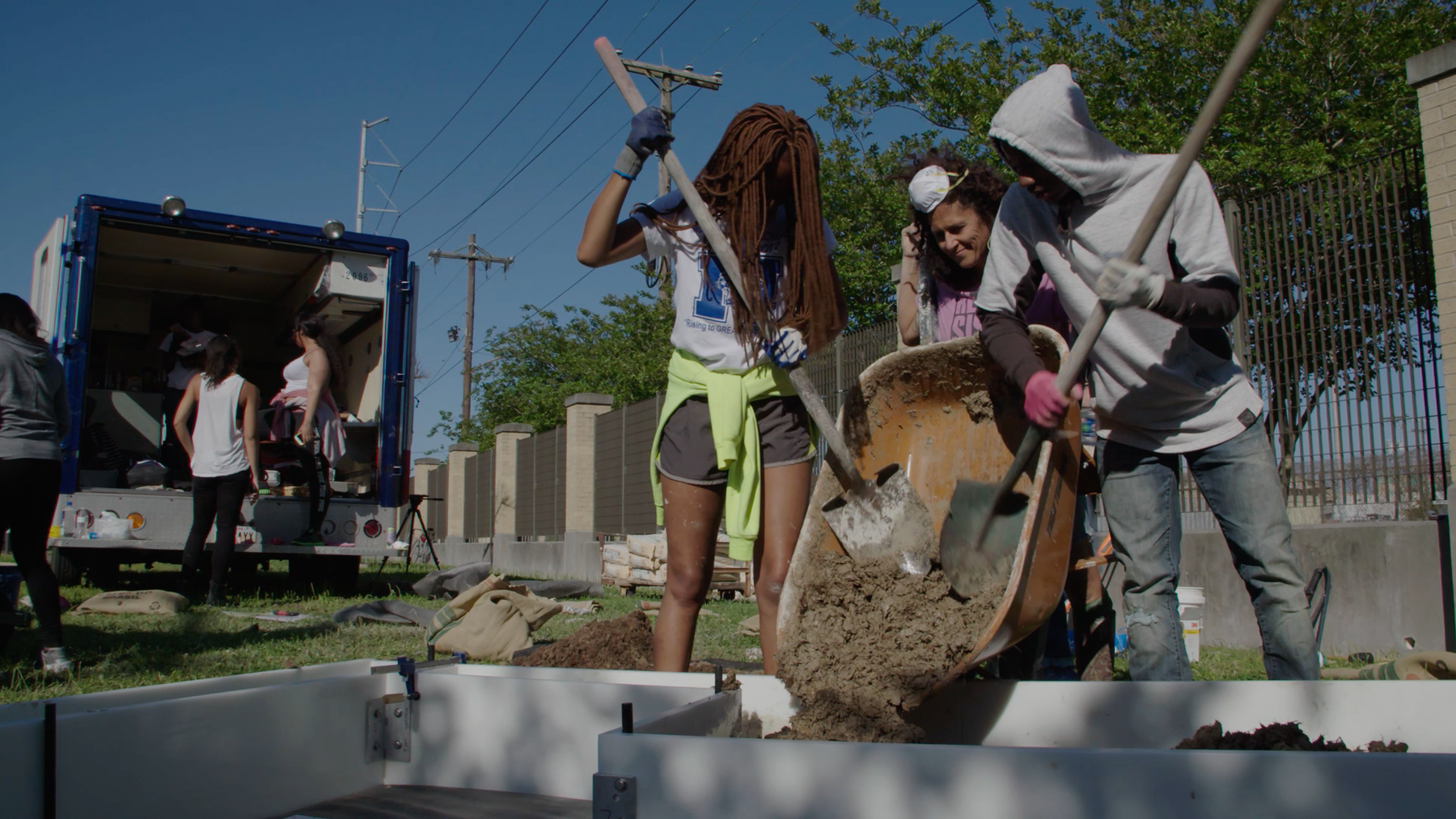 A woman stands between two young students who are shoveling dirt out of a wheelbarrow with two shovels into a garden bed made out of concrete.