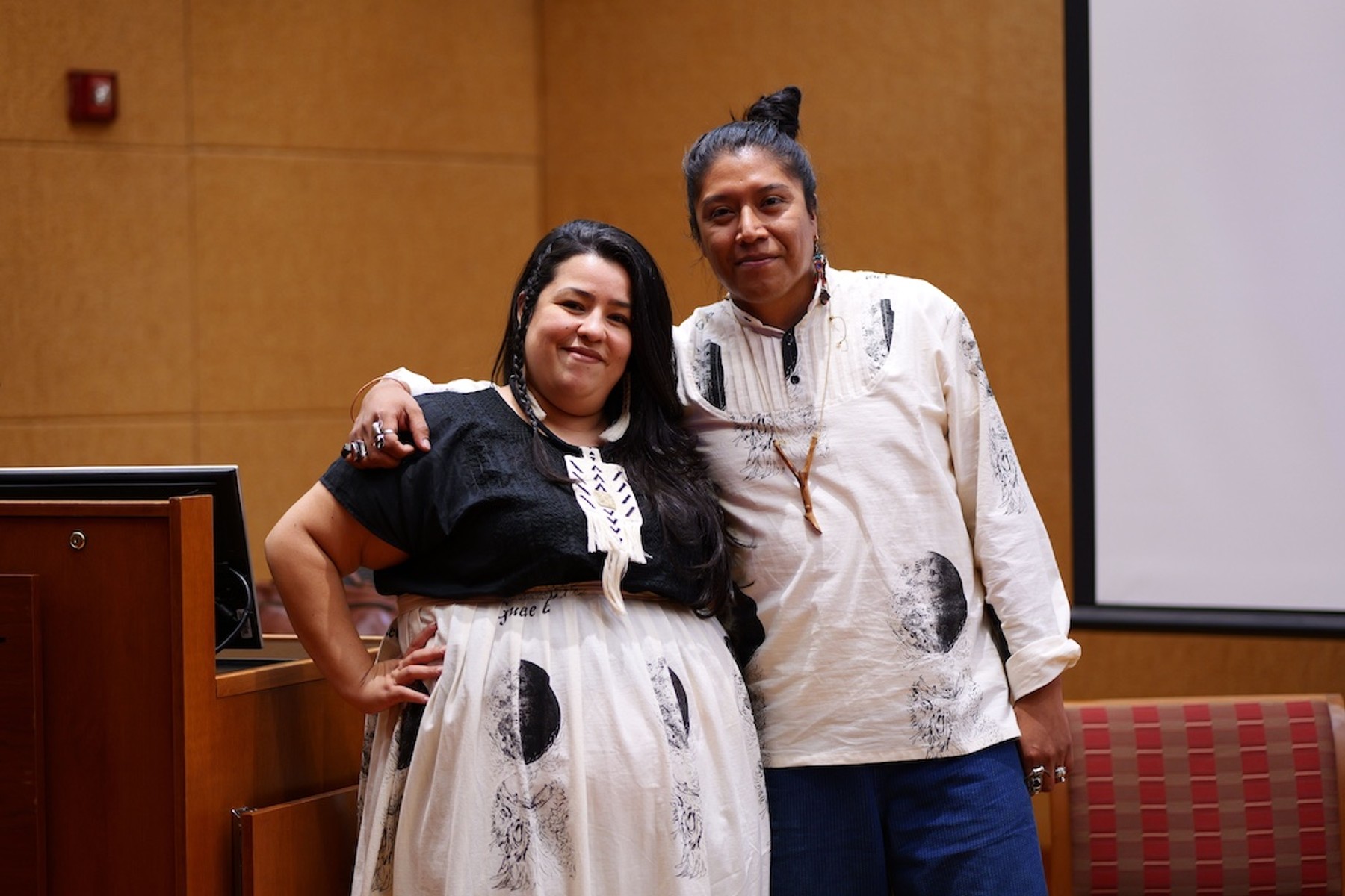 Erika Hirugami and Federico Cuatlacuatl stand in a warmly lit auditorium.
