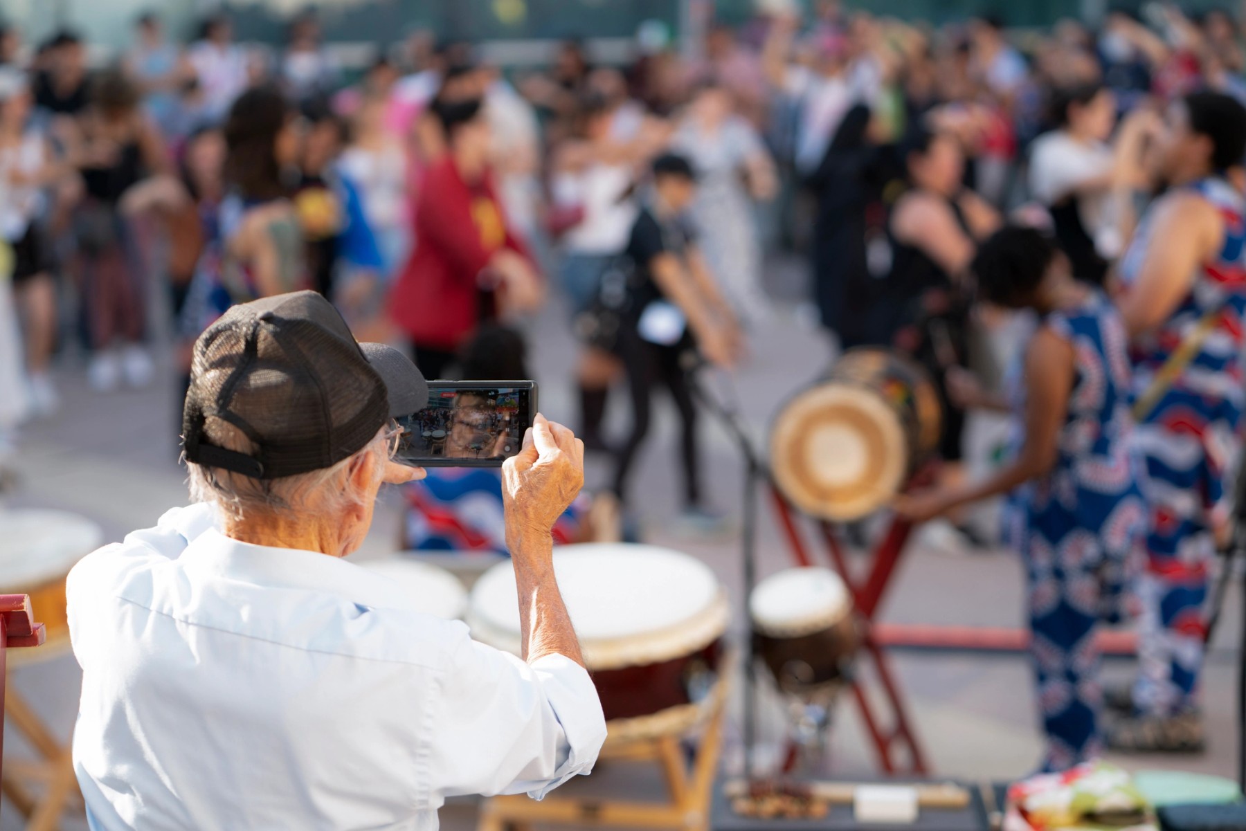 A Japanese American elder holds up his cellphone in the foreground to take a photo of the diverse group of people dancing in a circle. He wears a white shirt and black baseball cap. African drum musicians and their drums fill the middle of the frame. 