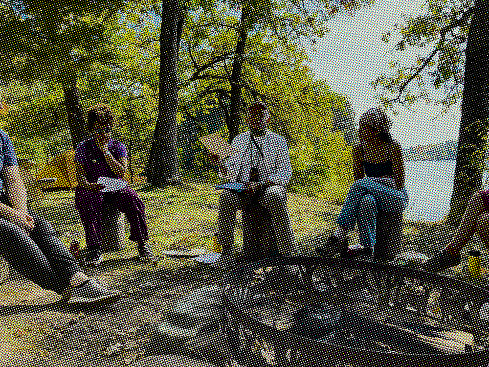 A group gathers around a firepit in the woods near a body of water during the day. 