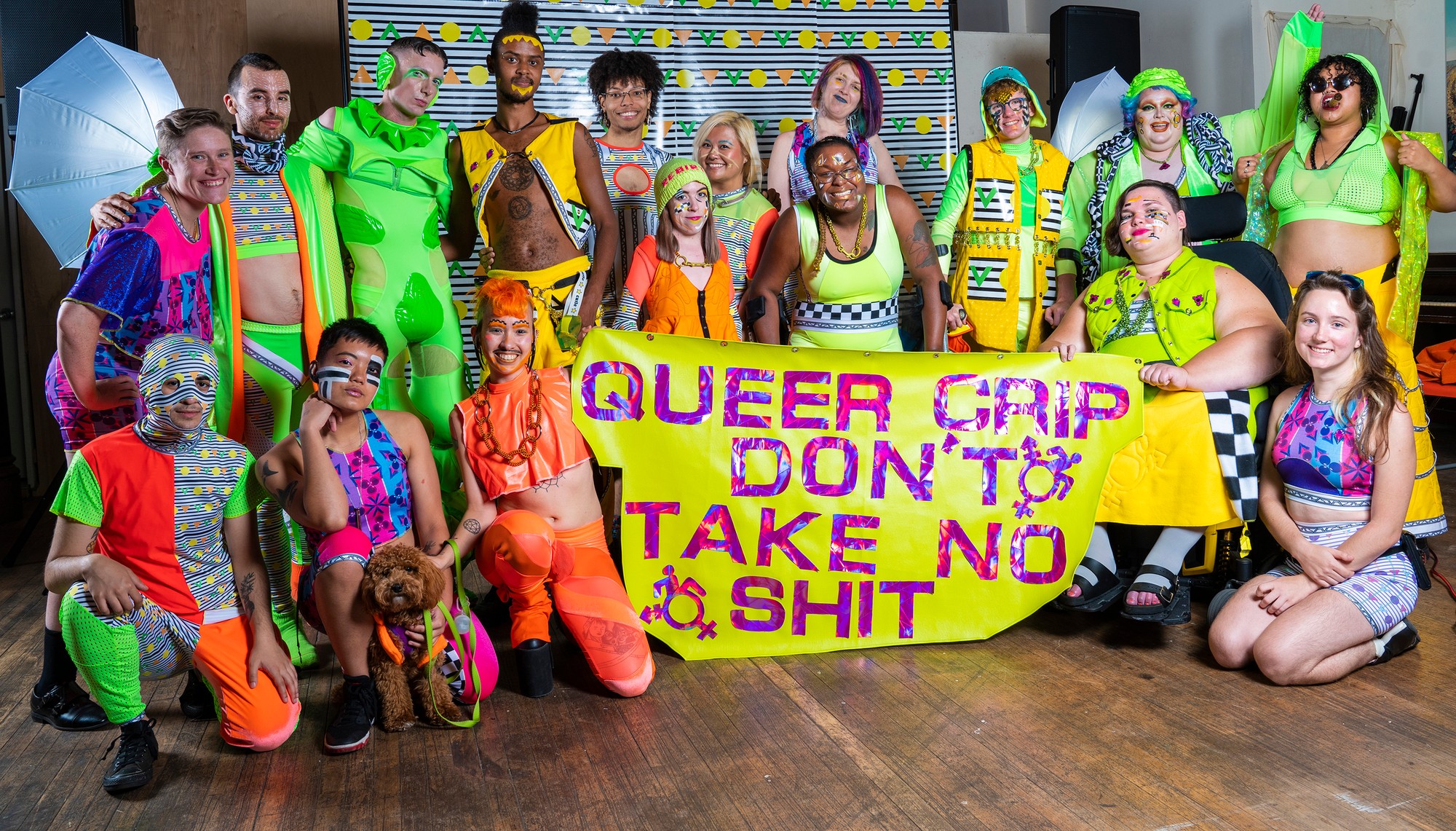 A group of seventeen people pose after a performance, wearing a collection comprised of neon orange, lime, and neon yellow fabrics with accents of black and white patterns. They pose with a yellow vinyl banner that says “Queercrip don’t take no sh*t” in holographic pink lettering, which references a line from videographer @TotallyQAF’s band EnGAYgement Party, featured in the first Radical Visibility Collective show. The banner is cut in a geometric asymmetrical blob shape and features two pink queercrip symbols.