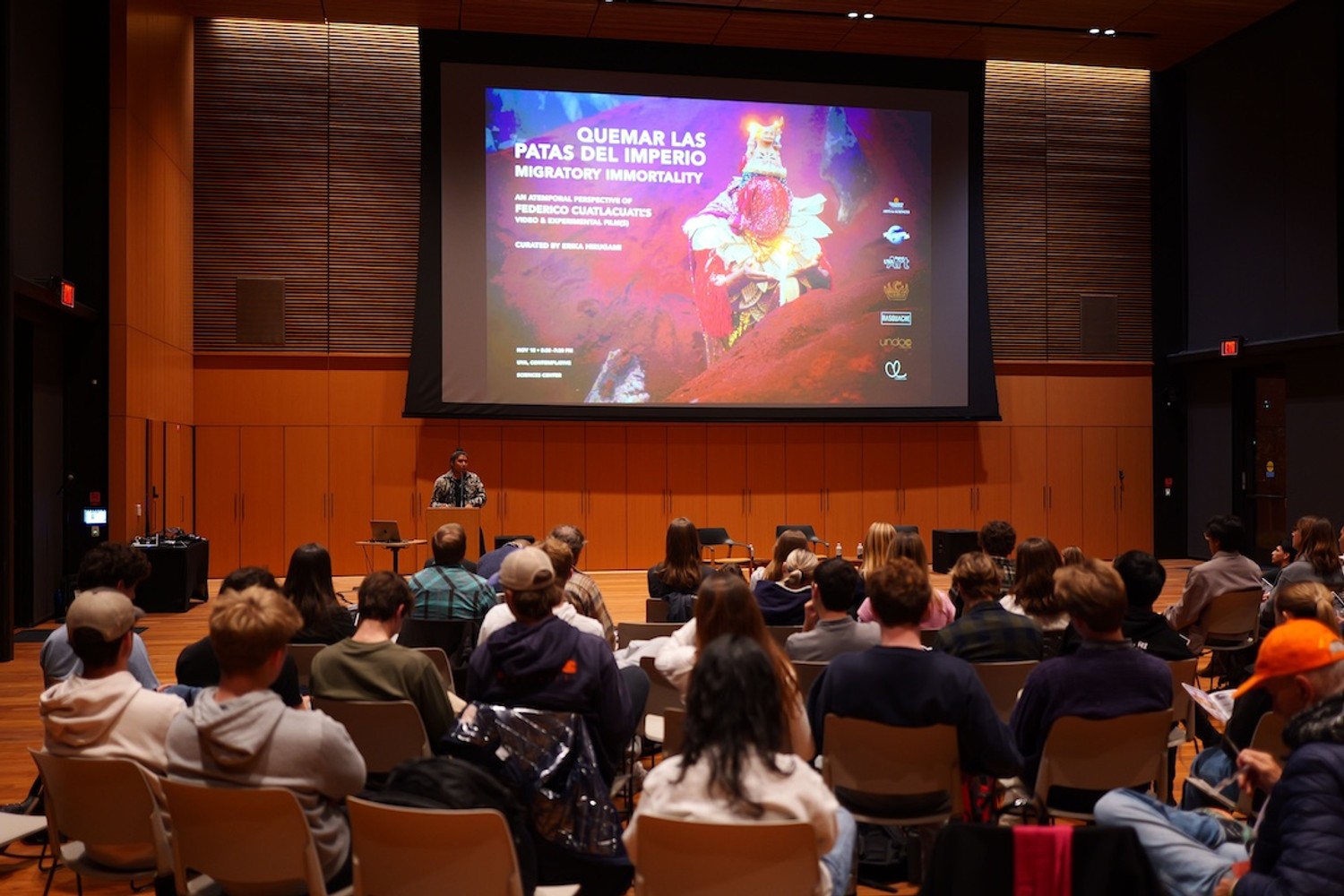 A crowded auditorium looks up at a projected video and a man lecturing below it.