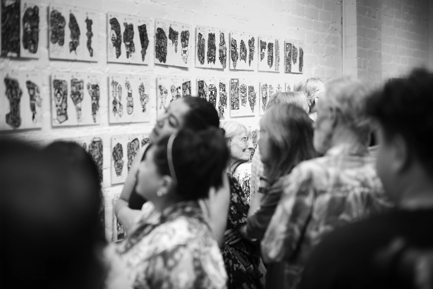 A black-and-white photograph of gallery visitors closely examining a grid of small, densely patterned ink drawings mounted in rows on a white brick wall. Several people lean in to look at the work, seen from behind and in profile.