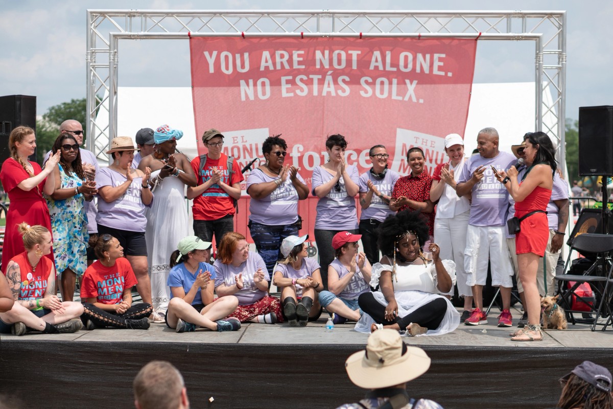 On an outdoor stage, people wearing colorful clothing applaud in front of a red banner with the phrase You Are Not Alone, No Estás Solx.