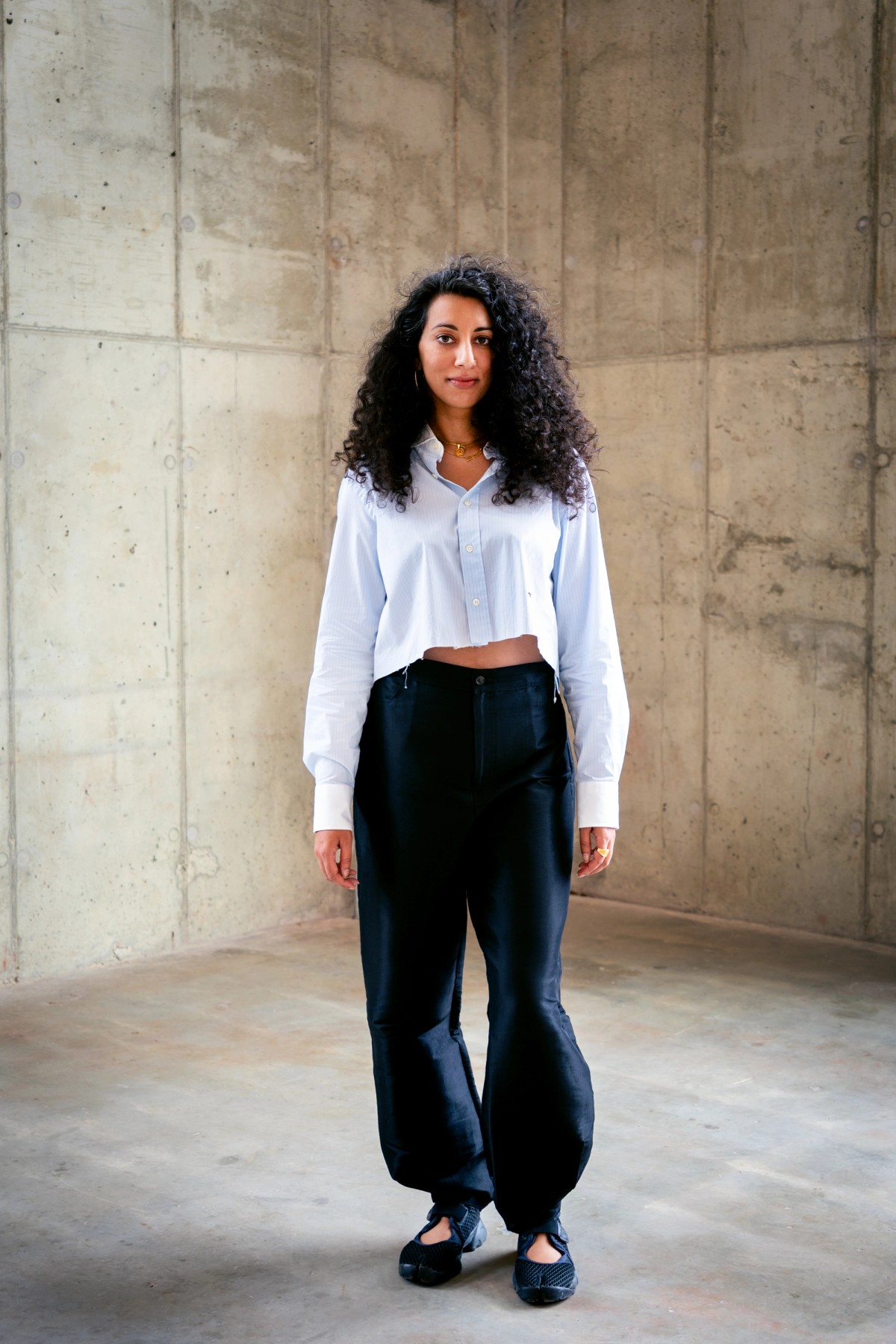 Diya is standing in the corner of the Powerhouse Arts Loft, a concrete warehouse-type building with natural light softly pouring in. She's Indian American, and her hair is big, black, and curly and flipped to one side. She's wearing a cropped button down, balloon black pants, and tabby sneakers. 