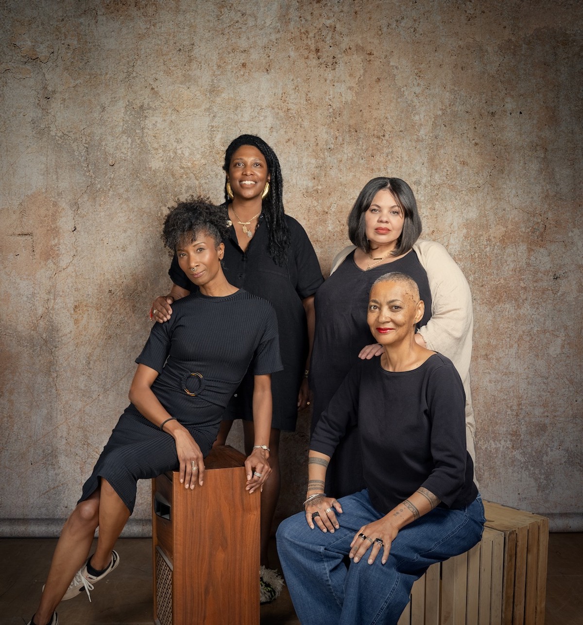 A studio portrait of four African American women dressed in black, posed together against a mottled brown backdrop. Two are seated on wooden crates in front, and two stand behind them, resting their hands on the others' shoulders. The composition is close and relaxed, conveying both individuality and collective presence.